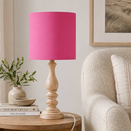 A table lamp with a cerise pink cylindrical shade and a turned light wood base, styled on a round wooden side table next to a beige armchair in a living room setting.