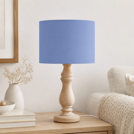 A table lamp with a cornflower blue cylindrical fabric shade and a natural wood turned base, shown on a wooden side table in a styled living room setting.