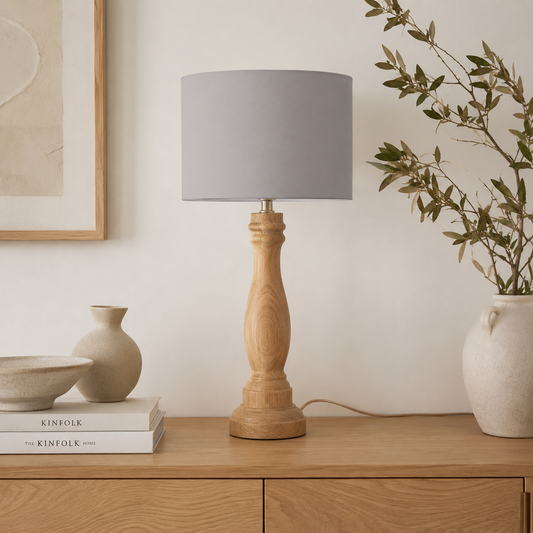 A table lamp with a light natural wood pedestal base and a grey fabric drum shade, styled on a wooden sideboard with books, a small vase, and a larger vase with dried branches against a white wall.