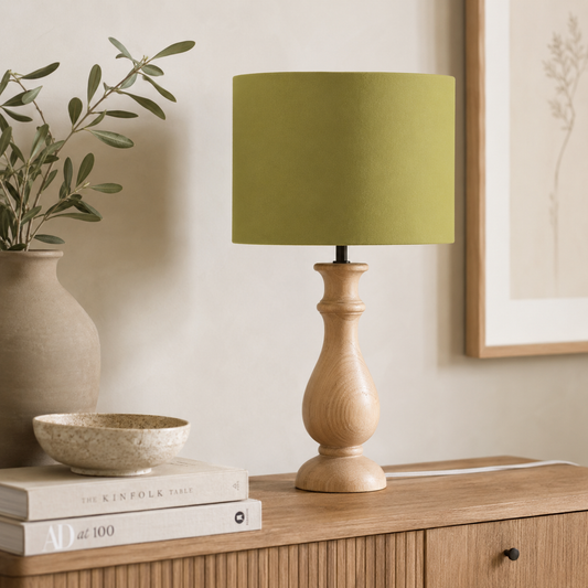 A table lamp with a muted olive green fabric drum shade and a light natural wood turned base, styled on a wooden sideboard next to a vase and books.