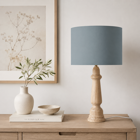 A table lamp with a petrol blue fabric drum shade and a turned natural wood pedestal base, styled on a light wood sideboard next to a white ceramic vase and a small bowl.