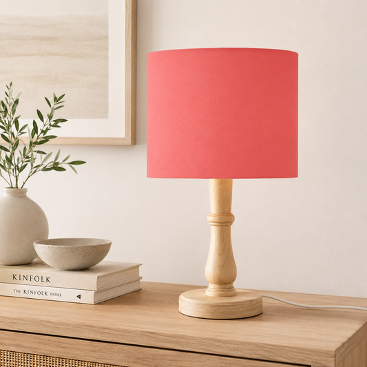A table lamp with a coral pink fabric drum shade and a natural light wood turned base, shown on a wooden console table in a styled room setting.