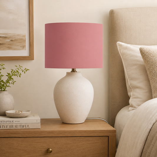 A table lamp with a fabric terracotta pink cylindrical shade and a textured off-white ceramic base, styled on a wooden nightstand next to a bed.