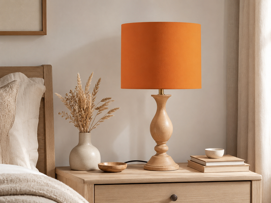 An orange drum lampshade table lamp with a turned light wood base, styled on a light wood nightstand next to a vase of dried grasses and books in a bedroom setting.