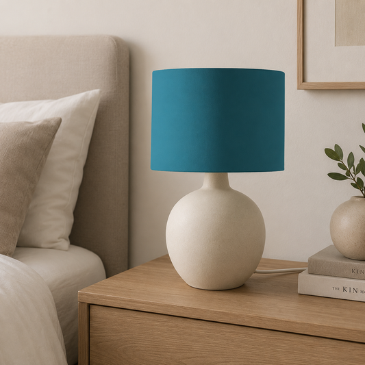 A table lamp with a matte teal blue drum shade and a textured cream-colored round base, styled on a wooden nightstand in a bedroom setting.