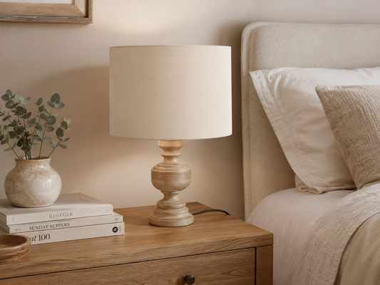 A rustic wooden table lamp with a beige fabric drum shade sits on a wooden nightstand next to a bed with neutral-toned bedding.