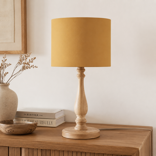 A table lamp with a warm mustard yellow fabric drum shade and a light natural wood turned base, shown on a wooden cabinet in a styled interior setting.