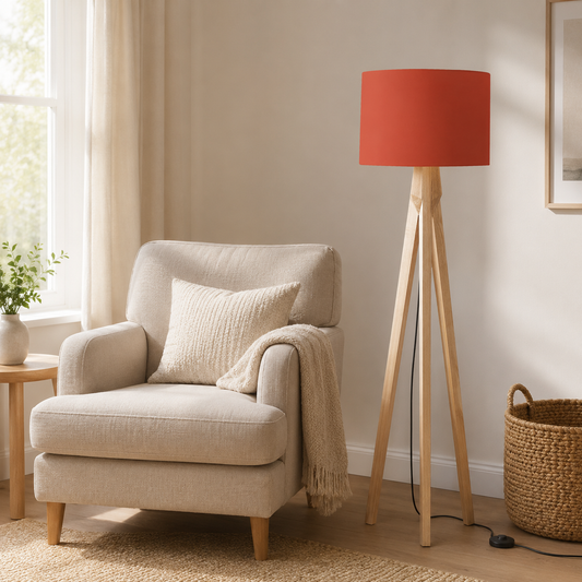 A modern floor lamp with a cylindrical Rust Red lampshade and three natural wood legs stands next to a beige armchair in a bright, neutral-toned living room.