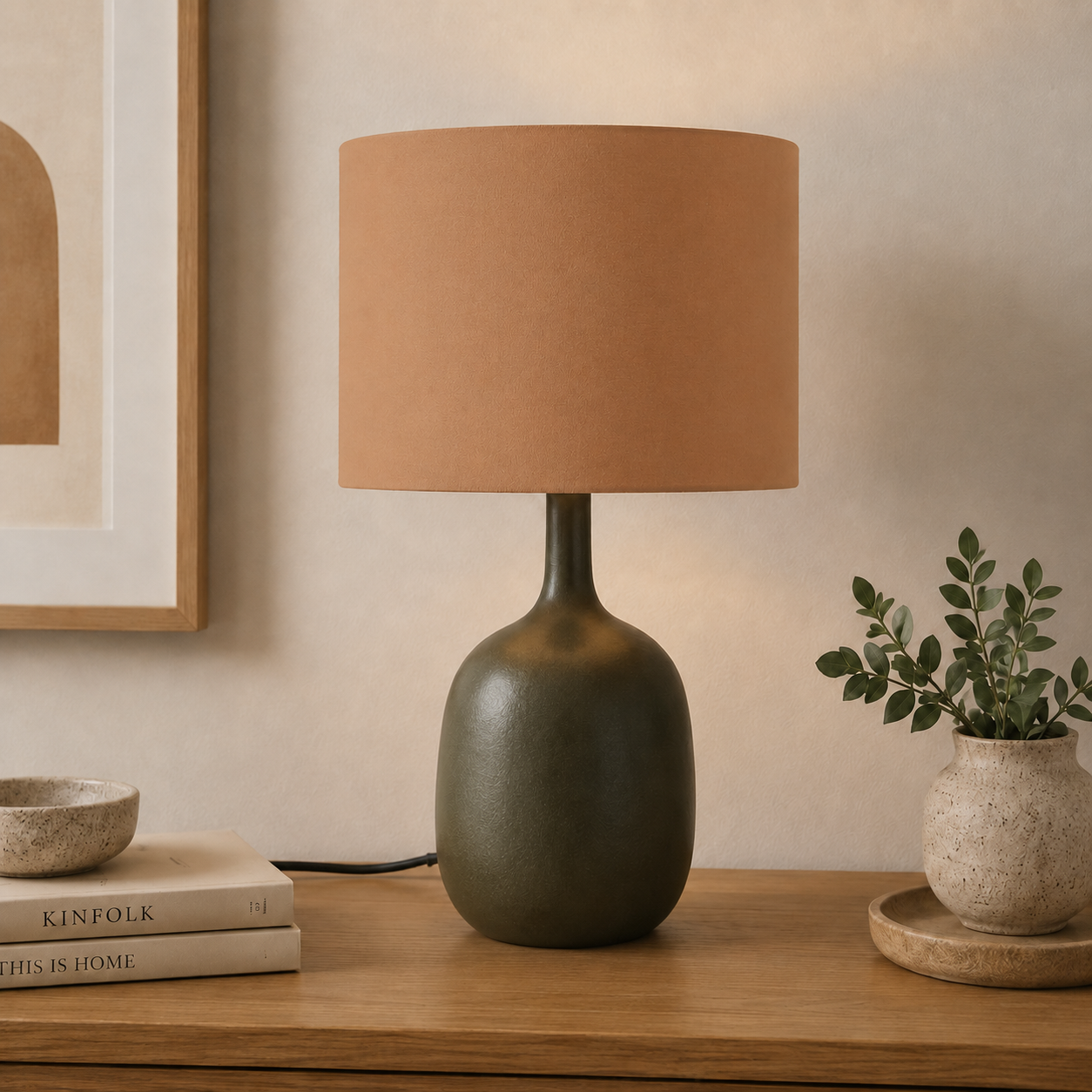 A table lamp with a matte olive green ceramic base and a warm toffee fabric drum shade, styled on a wooden console table with books and a small potted plant.
