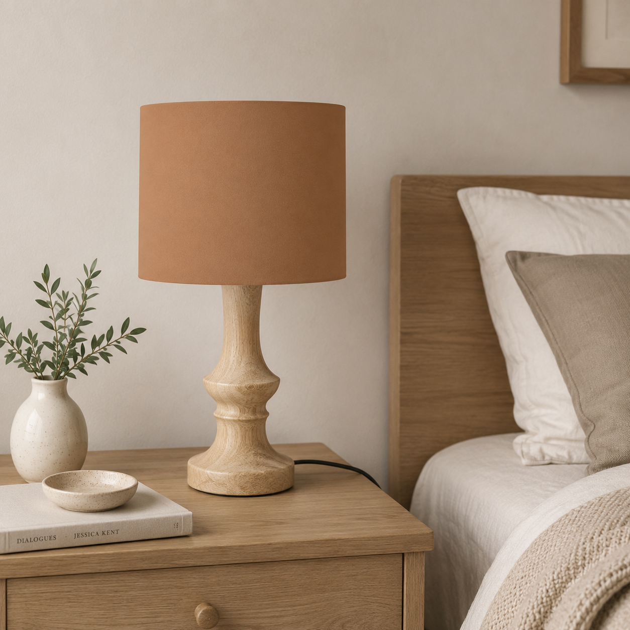 A table lamp with a warm toffee drum shade and a light natural wood pedestal base, styled on a wooden bedside table next to a bed.